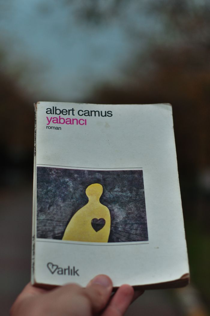 A hand holds an Albert Camus 'Yabanci' book, photographed outdoors.